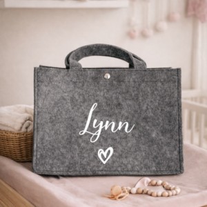 Grijze vilten tas met naam Lynn en hartje, ideaal kraamcadeau voor babykamer met knuffels op achtergrond